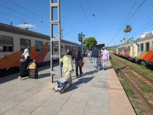 People walking along platform on a sunny day next to two railway carriages in Fes, Morocco.